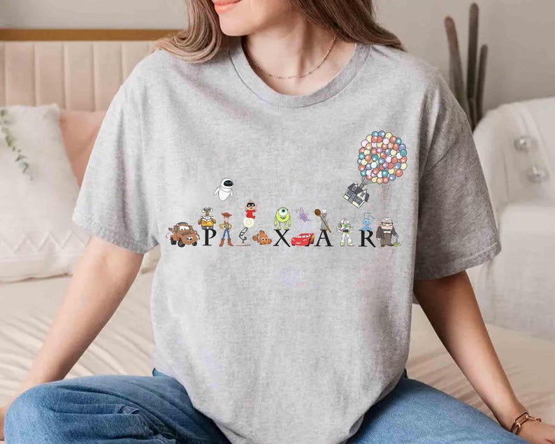 K&ouml;nnte beinhalten: Ein graues T-Shirt mit einem farbenfrohen Pixar-Design. Das Design zeigt verschiedene Charaktere, darunter Lightning McQueen und Wall-E, und das Wort "PIXAR". Eine Ansammlung von Ballons schwebt &uuml;ber dem "R" im Design.
