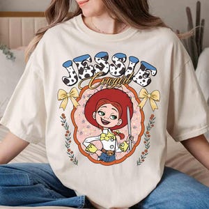 May include: A white t-shirt with a cartoon image of Jessie from Toy Story. The image is surrounded by a pink and yellow frame with the text "Jessie Cowgirl" in black and white.