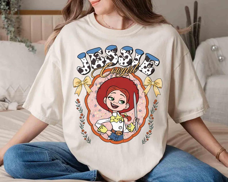 May include: A white t-shirt with a cartoon image of Jessie from Toy Story. The image is surrounded by a pink and yellow frame with the text "Jessie Cowgirl" in black and white.