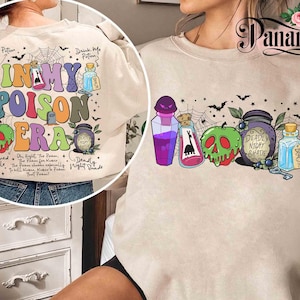 May include: Beige sweatshirt with a colorful graphic featuring potion bottles, a green skull apple, and the text "IN MY POISON ERA." The design includes bats, spiderwebs, and the words "Deadly Night Shade" and "Drink Me Potion."