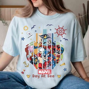 May include: Light blue t-shirt featuring a colorful graphic of Marvel characters and Disney characters. The design includes a ship's wheel, stars, and the text "MARVEL Day At Sea". The shirt is a casual, comfortable style.
