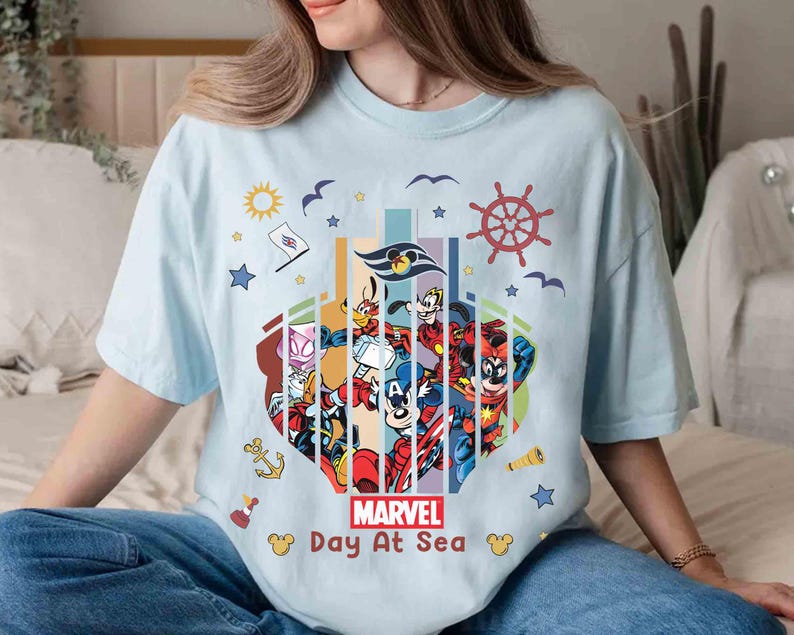 May include: Light blue t-shirt featuring a colorful graphic of Marvel characters and Disney characters. The design includes a ship's wheel, stars, and the text "MARVEL Day At Sea". The shirt is a casual, comfortable style.