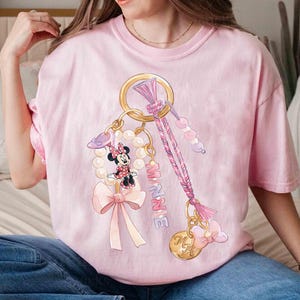 Op de afbeelding: Lichtroze T-shirt met een Minnie Mouse sleutelhanger ontwerp. Het ontwerp bevat een goudkleurige ring, een parelketting, een strik en de naam "MINNIE" in roze. Het shirt heeft een lichtroze kleur.