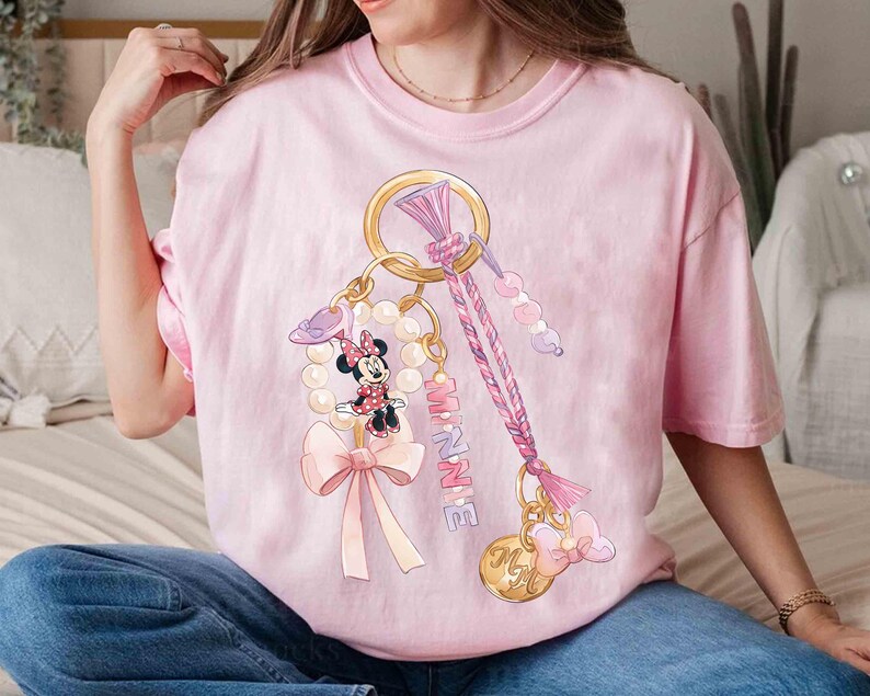 Op de afbeelding: Lichtroze T-shirt met een Minnie Mouse sleutelhanger ontwerp. Het ontwerp bevat een goudkleurige ring, een parelketting, een strik en de naam "MINNIE" in roze. Het shirt heeft een lichtroze kleur.