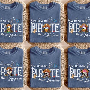 Peut inclure: Six t-shirts bleus avec un th&egrave;me de pirate. Chaque t-shirt pr&eacute;sente un personnage de dessin anim&eacute; diff&eacute;rent portant un chapeau de pirate, le mot "PIRATE" et la phrase "Life for me". Les t-shirts sont dispos&eacute;s sur une surface tiss&eacute;e.