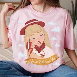 May include: A light pink t-shirt featuring a graphic illustration of a blonde person wearing a red hat and holding a small doll. The design includes white clouds and a yellow banner at the bottom with the text I Knew It, I Knew You.