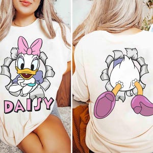Puede incluir: Camiseta color crema con un dibujo animado de Daisy Duck con un lazo rosa, un top morado y el nombre "DAISY" en rosa. La parte trasera de la camiseta muestra las piernas y los pies de Daisy. Una camiseta divertida e informal.