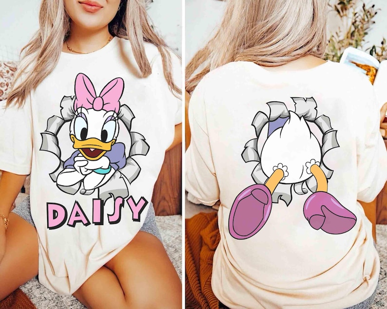 Puede incluir: Camiseta color crema con un dibujo animado de Daisy Duck con un lazo rosa, un top morado y el nombre "DAISY" en rosa. La parte trasera de la camiseta muestra las piernas y los pies de Daisy. Una camiseta divertida e informal.