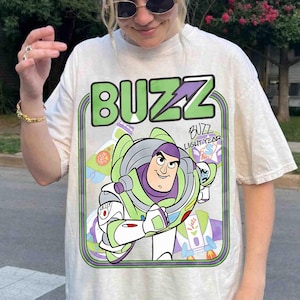 May include: A cream-colored short-sleeve t-shirt featuring a large graphic print of the character Buzz Lightyear. The design includes the text BUZZ in large green letters at the top and the name Buzz Lightyear written in a stylized script font.