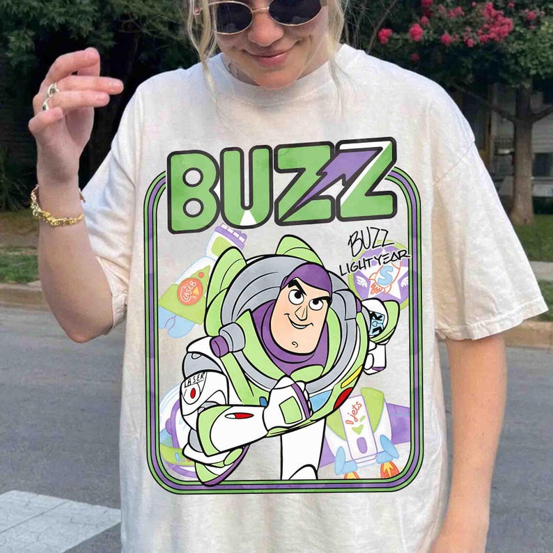 May include: A cream-colored short-sleeve t-shirt featuring a large graphic print of the character Buzz Lightyear. The design includes the text BUZZ in large green letters at the top and the name Buzz Lightyear written in a stylized script font.
