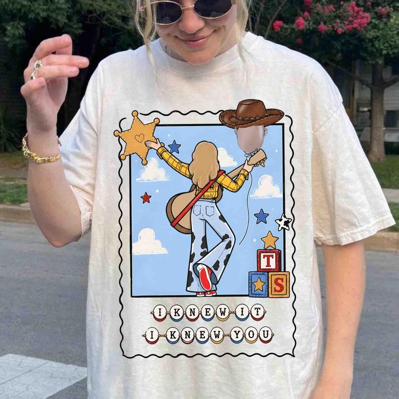 May include: A cream-colored t-shirt featuring a graphic print of a cartoon character with blonde hair, a yellow plaid shirt, and cow-print pants, holding a guitar and a sheriff star. The text reads I KNEW IT I KNEW YOU with toy blocks.