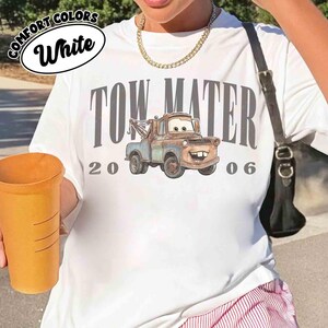 May include: White t-shirt featuring a graphic of Tow Mater from the movie Cars, with the text "TOW MATER 2006". The shirt has a round neckline and short sleeves. The words "COMFORT COLORS" and "White" are printed on the top left.