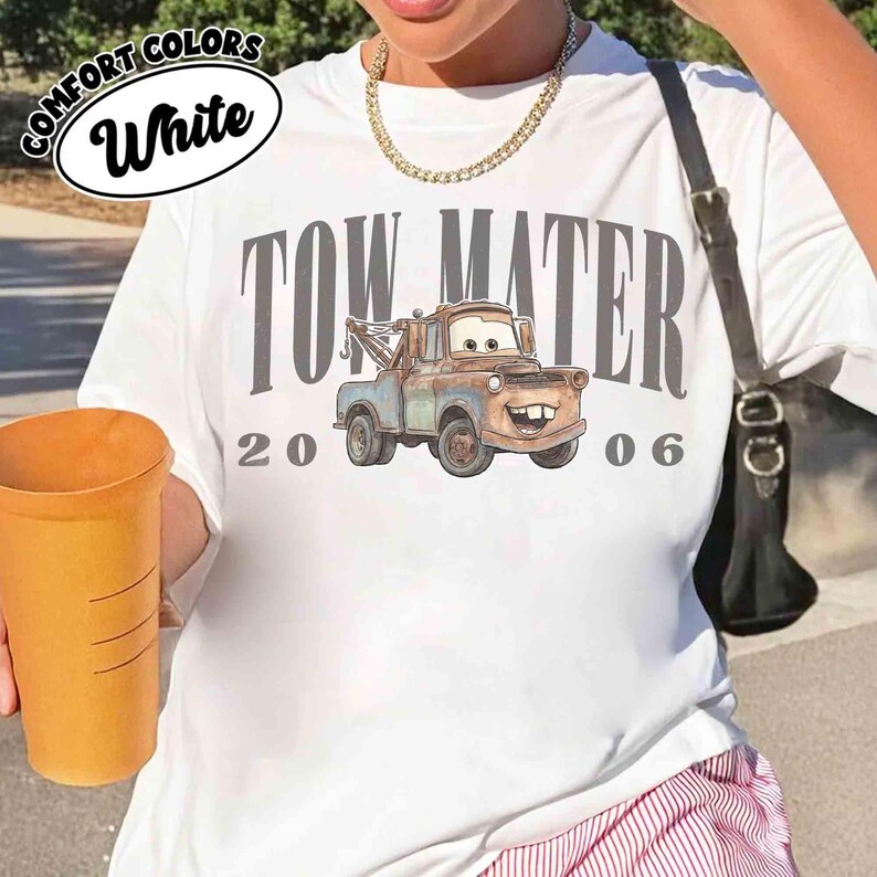 May include: White t-shirt featuring a graphic of Tow Mater from the movie Cars, with the text "TOW MATER 2006". The shirt has a round neckline and short sleeves. The words "COMFORT COLORS" and "White" are printed on the top left.