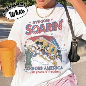 May include: White t-shirt with a graphic featuring Mickey Mouse, Goofy, and Donald Duck flying over the Statue of Liberty. The shirt has the text "SOARIN'" and "ACROSS AMERICA 250 years of freedom" in red and blue.