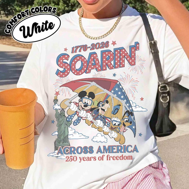 May include: White t-shirt with a graphic featuring Mickey Mouse, Goofy, and Donald Duck flying over the Statue of Liberty. The shirt has the text "SOARIN'" and "ACROSS AMERICA 250 years of freedom" in red and blue.