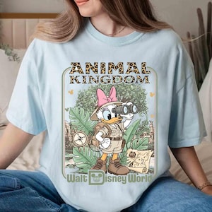 Puede incluir: Camiseta azul claro con un dise&ntilde;o de Daisy Duck con ropa de safari, sosteniendo unos prism&aacute;ticos. El dise&ntilde;o incluye el texto "ANIMAL KINGDOM" en estampado de leopardo y "Walt Disney World". La camiseta tiene mangas cortas y cuello redondo.