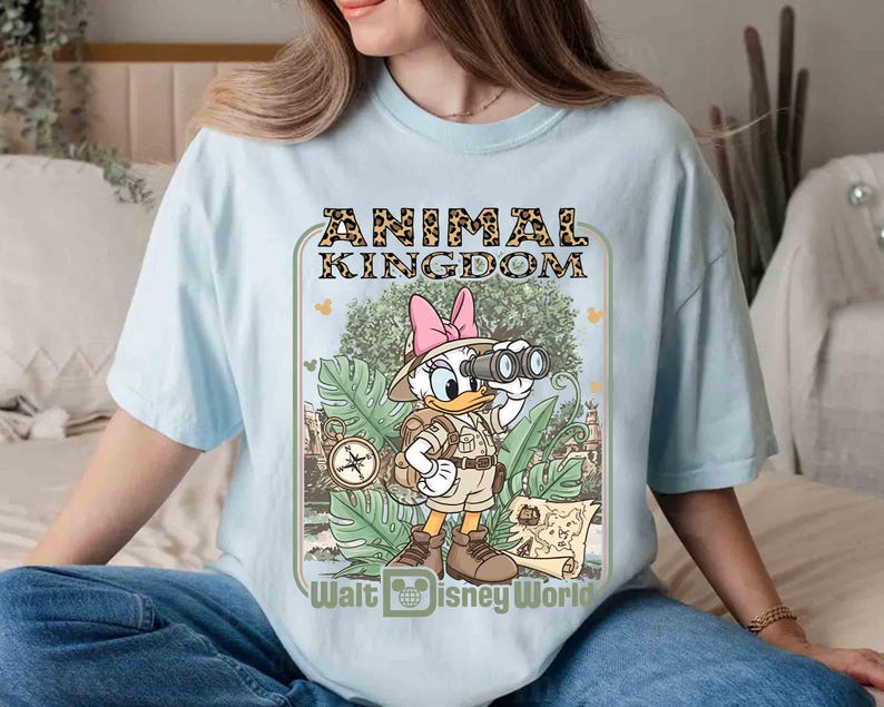 Puede incluir: Camiseta azul claro con un dise&ntilde;o de Daisy Duck con ropa de safari, sosteniendo unos prism&aacute;ticos. El dise&ntilde;o incluye el texto "ANIMAL KINGDOM" en estampado de leopardo y "Walt Disney World". La camiseta tiene mangas cortas y cuello redondo.