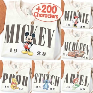 May include: Cream-colored t-shirts featuring various Disney characters: Mickey Mouse, Minnie Mouse, Lightning McQueen, Winnie the Pooh, Stitch, and Ariel. Each shirt has the character's name and a year printed on it.