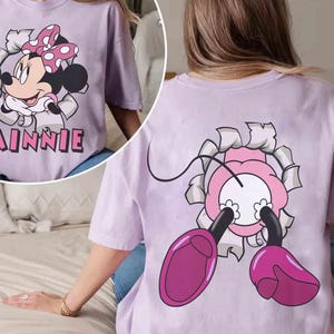 May include: A light purple t-shirt featuring a Minnie Mouse graphic. The front of the shirt shows Minnie breaking through a torn-paper effect, with the name "Minnie" below. The back of the shirt shows Minnie's legs and part of her head.