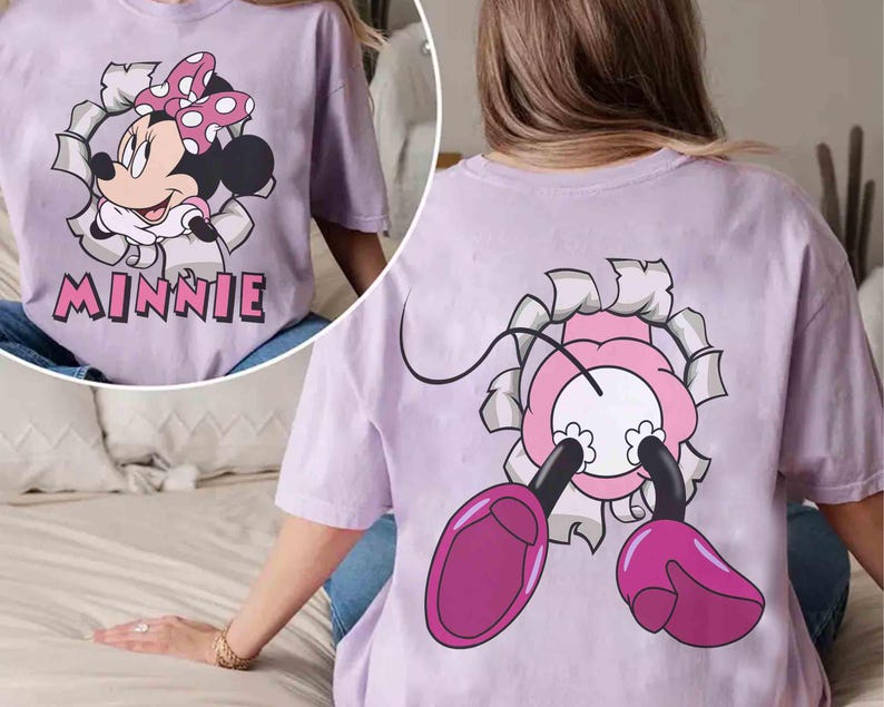 May include: A light purple t-shirt featuring a Minnie Mouse graphic. The front of the shirt shows Minnie breaking through a torn-paper effect, with the name "Minnie" below. The back of the shirt shows Minnie's legs and part of her head.