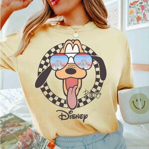 May include: A yellow t-shirt with a cartoon image of Pluto wearing sunglasses. The sunglasses reflect an image of Cinderella's castle. The text "Pluto" and "Disney" are printed on the shirt.