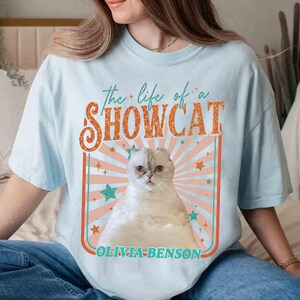 Może przedstawiać: Jasnoniebieska koszulka z napisem "the life of a SHOWCAT" w kolorze pomarańczowym i białym, z ilustracją kota. Kot jest biały ze złożonymi uszami. Pod ilustracją znajduje się napis "OLIVIA-BENSON".