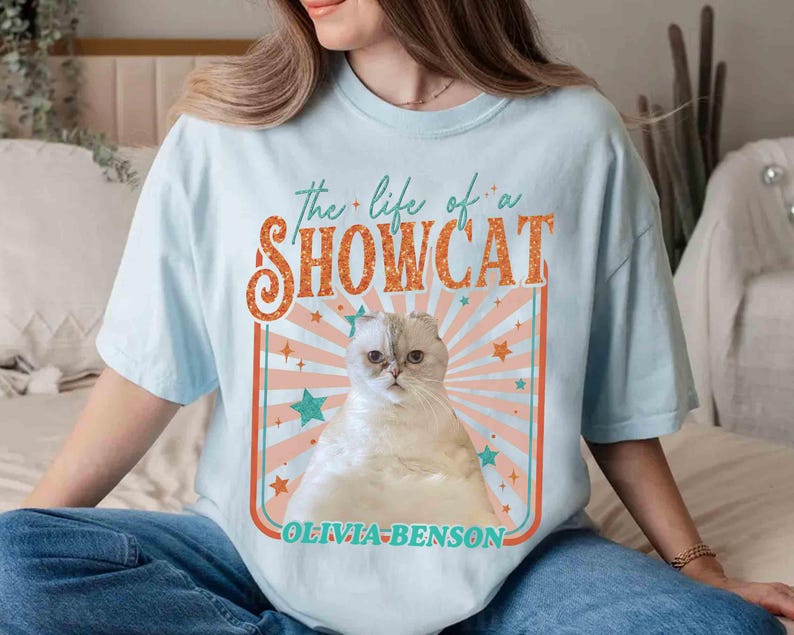 Może przedstawiać: Jasnoniebieska koszulka z napisem "the life of a SHOWCAT" w kolorze pomarańczowym i białym, z ilustracją kota. Kot jest biały ze złożonymi uszami. Pod ilustracją znajduje się napis "OLIVIA-BENSON".