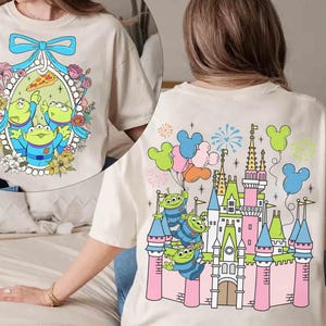 May include: A white t-shirt featuring a cartoon image of three green aliens from the Toy Story franchise in front of a pink castle with fireworks. The castle is reminiscent of a famous theme park.