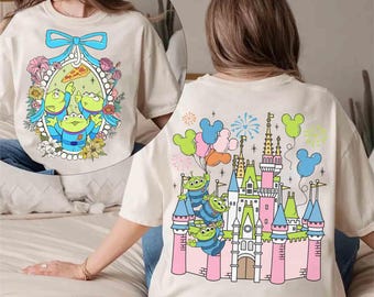Puede incluir: Una camiseta blanca con una imagen de dibujos animados de tres extraterrestres verdes de la franquicia Toy Story frente a un castillo rosa con fuegos artificiales. El castillo recuerda a un famoso parque de atracciones.