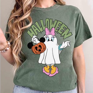 May include: Green t-shirt with a Halloween graphic featuring a cartoon character dressed as a ghost, holding a Mickey Mouse pumpkin. The word "HALLOWEEN" is printed above the character in green letters. The character has a pink bow and yellow shoes.