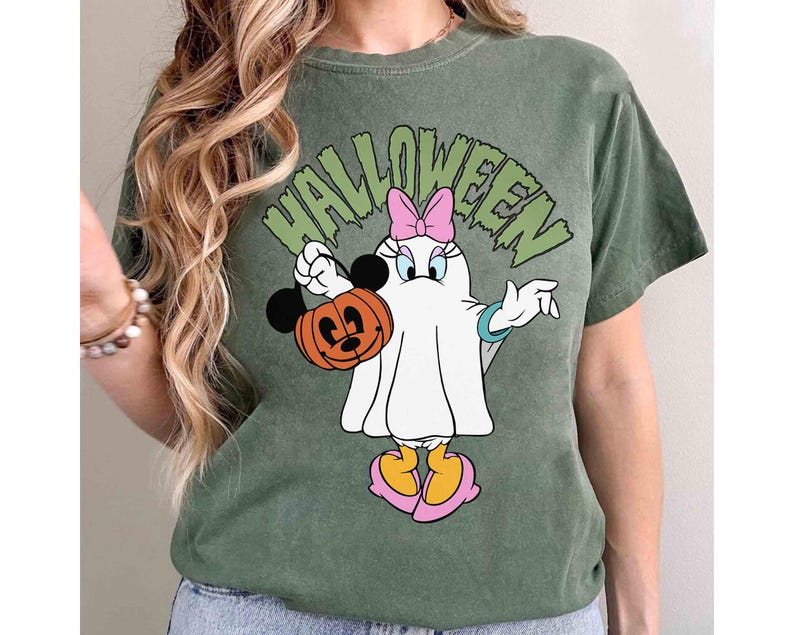 May include: Green t-shirt with a Halloween graphic featuring a cartoon character dressed as a ghost, holding a Mickey Mouse pumpkin. The word "HALLOWEEN" is printed above the character in green letters. The character has a pink bow and yellow shoes.