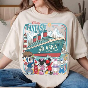 May include: Cream-colored t-shirt featuring a colorful graphic of a cruise ship with the text "Alaska" and "Disney Fantasy." The design includes cartoon characters and a lighthouse. The shirt has a relaxed fit.
