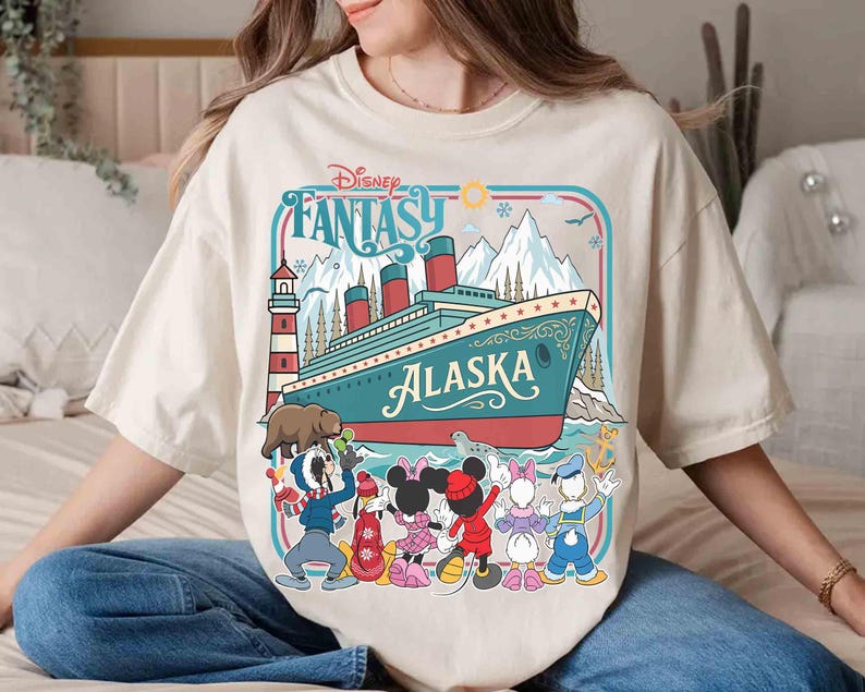 May include: Cream-colored t-shirt featuring a colorful graphic of a cruise ship with the text "Alaska" and "Disney Fantasy." The design includes cartoon characters and a lighthouse. The shirt has a relaxed fit.