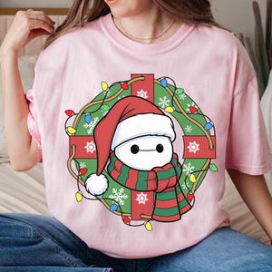 Op de afbeelding: Lichtroze T-shirt met een stripfiguur met een kerstmuts en een rood-groene sjaal, omringd door een kerstkrans met lichtjes en rode cadeauverpakkingen. Het ontwerp heeft een feestelijk, vakantiethema.