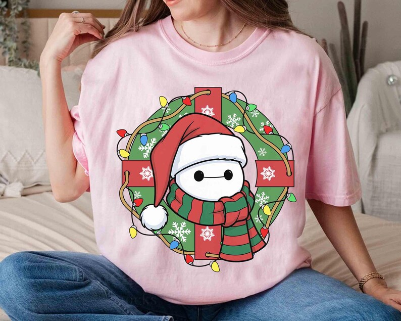 Op de afbeelding: Lichtroze T-shirt met een stripfiguur met een kerstmuts en een rood-groene sjaal, omringd door een kerstkrans met lichtjes en rode cadeauverpakkingen. Het ontwerp heeft een feestelijk, vakantiethema.