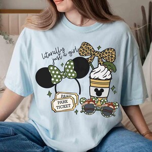 May include: Light blue t-shirt with a graphic featuring Minnie Mouse ears, a coffee cup, sunglasses, and park tickets. The text "literally just a girl" is above the Minnie Mouse ears. The shirt has a relaxed fit.