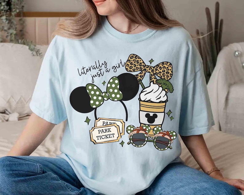 May include: Light blue t-shirt with a graphic featuring Minnie Mouse ears, a coffee cup, sunglasses, and park tickets. The text "literally just a girl" is above the Minnie Mouse ears. The shirt has a relaxed fit.