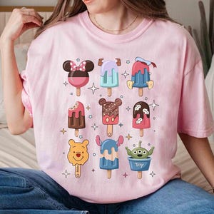 May include: A light pink t-shirt featuring colorful cartoon popsicle designs. The popsicles have character-themed designs, including Minnie Mouse, Mickey Mouse, and Winnie the Pooh. The shirt is a crew neck and has short sleeves.