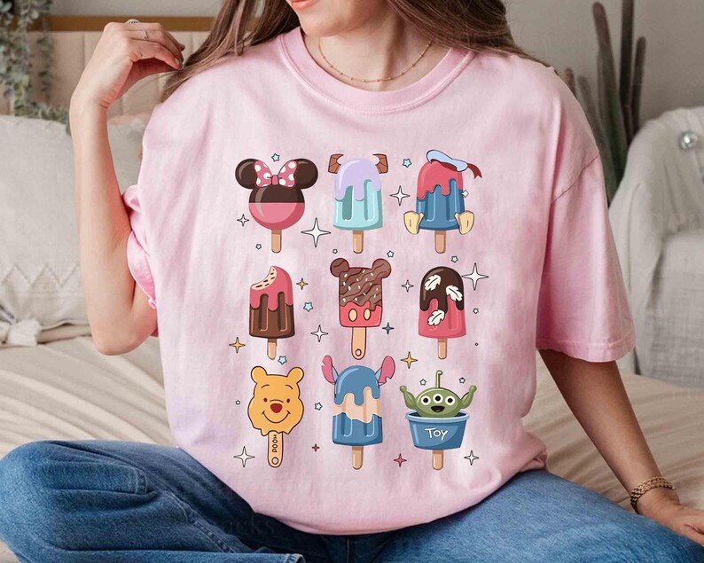 May include: A light pink t-shirt featuring colorful cartoon popsicle designs. The popsicles have character-themed designs, including Minnie Mouse, Mickey Mouse, and Winnie the Pooh. The shirt is a crew neck and has short sleeves.