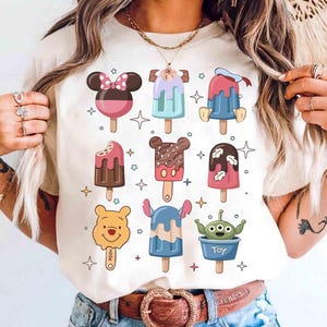 May include: Cream-colored t-shirt featuring colorful cartoon popsicle designs. The popsicles have character-themed designs, including Mickey Mouse, Donald Duck, and Winnie the Pooh. The word "Toy" is printed on one of the popsicles.