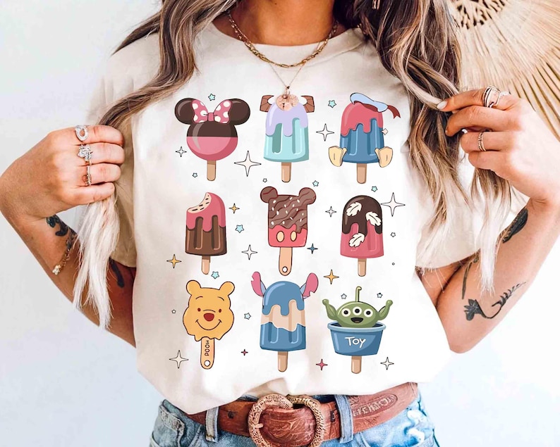 May include: Cream-colored t-shirt featuring colorful cartoon popsicle designs. The popsicles have character-themed designs, including Mickey Mouse, Donald Duck, and Winnie the Pooh. The word "Toy" is printed on one of the popsicles.