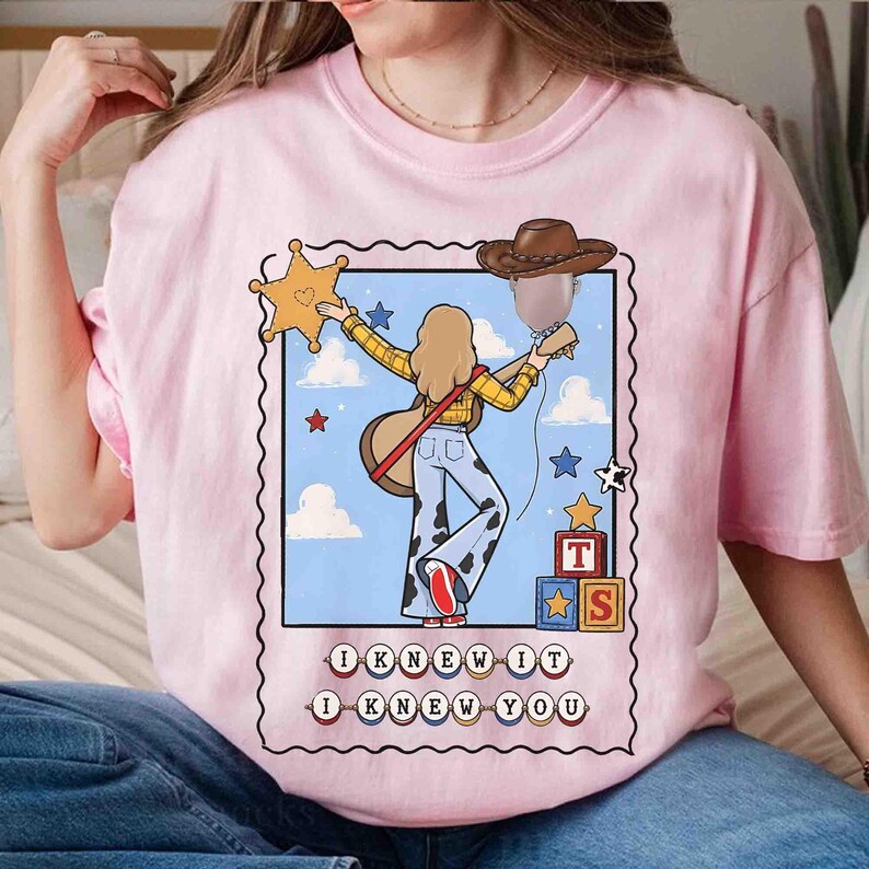 May include: A light pink t-shirt featuring a graphic illustration of a person with long hair wearing a yellow plaid shirt and cow-print pants, holding a guitar. The design includes a brown cowboy hat, a gold sheriff star, and blocks with the letters T and S. The text reads I KNEW IT I KNEW YOU.