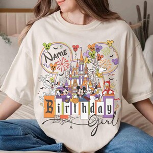 May include: Cream-colored t-shirt featuring a colorful graphic of a castle, balloons, and cartoon characters. The shirt has the words "Name" and "Birthday Girl" printed on it. The design includes fireworks and spiderwebs.