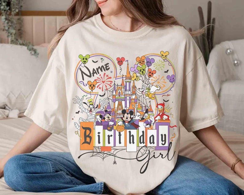 May include: Cream-colored t-shirt featuring a colorful graphic of a castle, balloons, and cartoon characters. The shirt has the words "Name" and "Birthday Girl" printed on it. The design includes fireworks and spiderwebs.