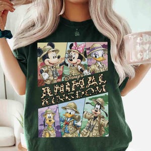 K&ouml;nnte beinhalten: Dunkelgr&uuml;nes T-Shirt mit einer Grafik der Disney-Figuren Mickey Mouse, Minnie Mouse, Donald Duck, Daisy Duck, Goofy und Pluto in Safari-Outfits. Der Text "Disney's Animal Kingdom" ist ebenfalls abgebildet.