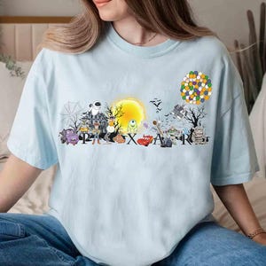 May include: Light blue t-shirt featuring a colorful graphic of various Pixar characters and the word "PIXAR". The design includes characters from Monsters, Inc., Toy Story, and Cars, among others. A yellow sun and a cluster of balloons are also featured.