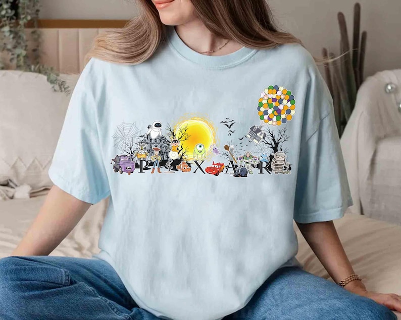 May include: Light blue t-shirt featuring a colorful graphic of various Pixar characters and the word "PIXAR". The design includes characters from Monsters, Inc., Toy Story, and Cars, among others. A yellow sun and a cluster of balloons are also featured.