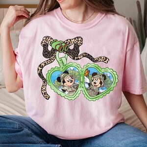 Puede incluir: Camiseta rosa con un gr&aacute;fico de Mickey Mouse y Minnie Mouse con sombreros de safari y sosteniendo binoculares. El gr&aacute;fico est&aacute; rodeado por un dise&ntilde;o de estampado de leopardo.
