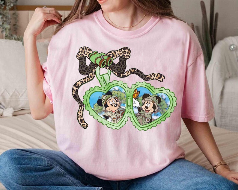 Puede incluir: Camiseta rosa con un gr&aacute;fico de Mickey Mouse y Minnie Mouse con sombreros de safari y sosteniendo binoculares. El gr&aacute;fico est&aacute; rodeado por un dise&ntilde;o de estampado de leopardo.
