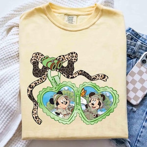 Puede incluir: Una camiseta amarilla con un gr&aacute;fico de dos personajes de dibujos animados en un medall&oacute;n en forma de coraz&oacute;n. El medall&oacute;n est&aacute; rodeado por una cinta de estampado de leopardo marr&oacute;n y negro. Los personajes llevan atuendos de safari y sostienen binoculares. El texto "COMFORT COLORS" es visible en la camiseta.